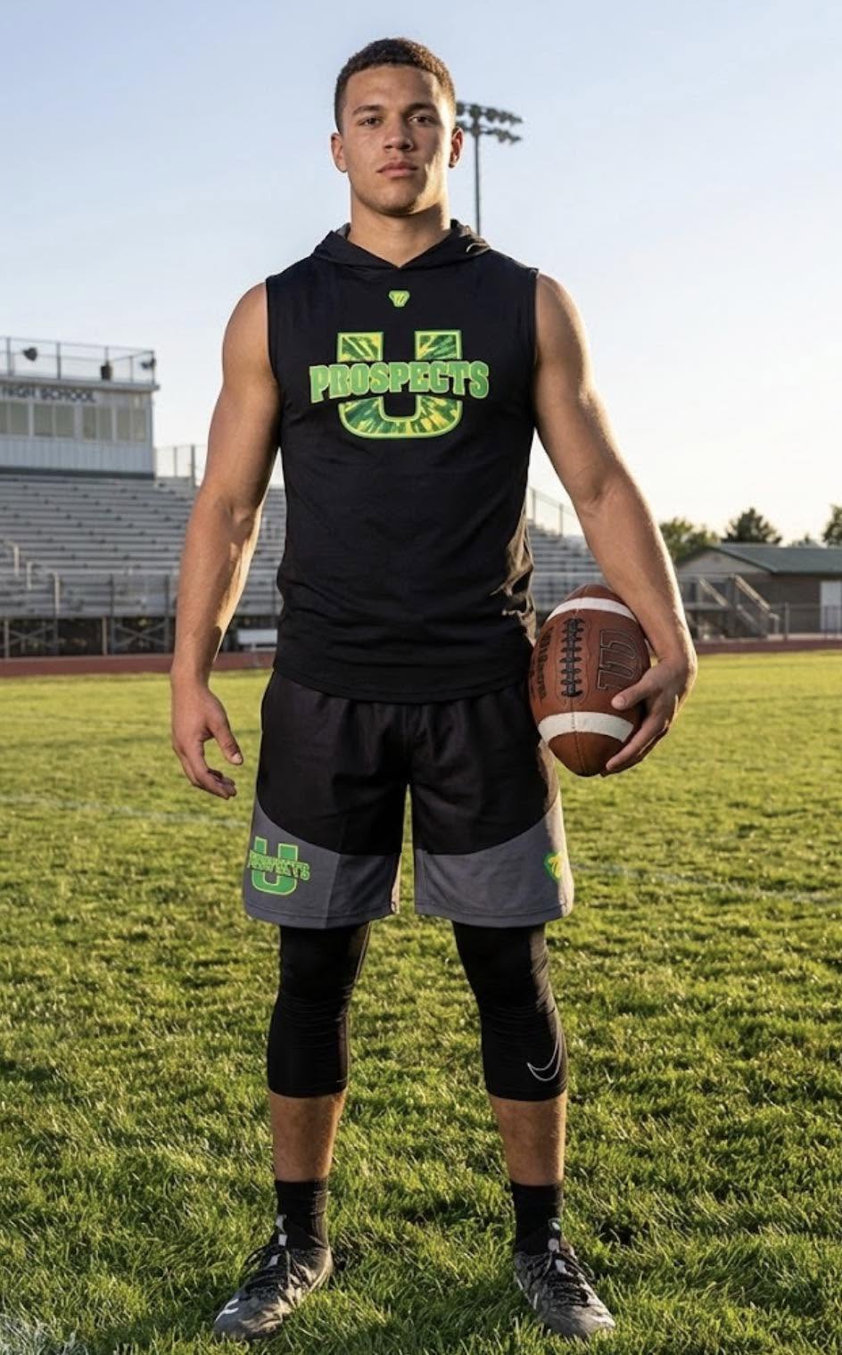 A Utah Prospects player in a black U Prospects sleeveless hoodie holding a football on the field at golden hour